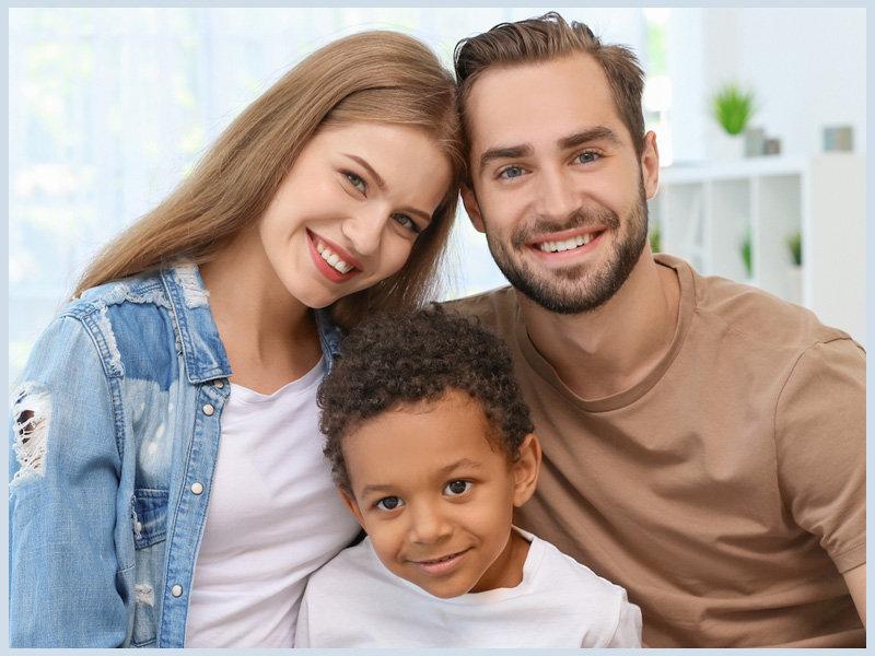 Smiling family of three, embracing in a bright room. Woman in blue denim, child in white, man in tan. Joyful, cozy atmosphere.